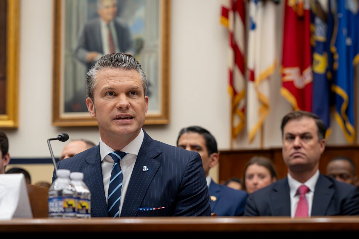 Defense Secretary Pete Hegseth speaks while seated at a table with a microphone attached, and a seated audience behind him.