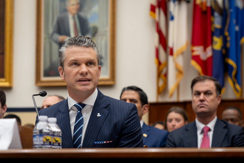 Defense Secretary Pete Hegseth speaks while seated at a table with a microphone attached, and a seated audience behind him.