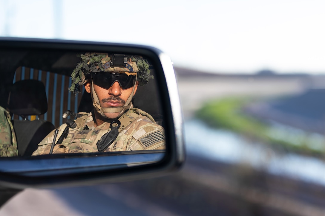 A soldier's reflection is seen in the rearview mirror of a vehicle as he looks out the window toward a blurred background.
