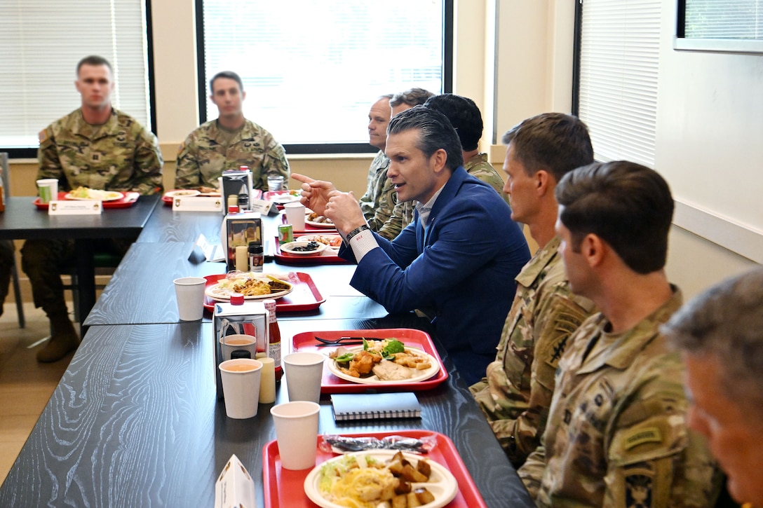 A man in a suit sits with a tray of food in  a room at a table with soldiers also eating food.