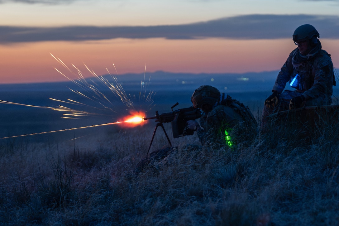 Sparks fly as a guardsman fires a weapon while lying on a hill as a fellow guardsman watches under a gray, pinkish sky.