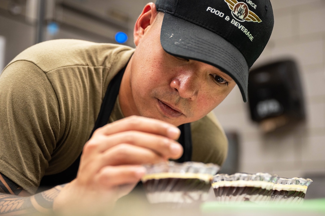 A close-up of an airman in an apron sprinkling chocolate on top of a dessert.