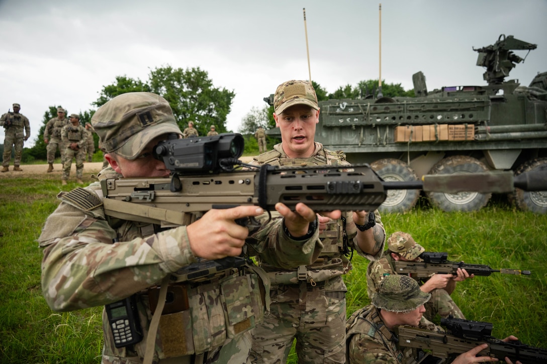 A soldier guides another soldier as they aim a weapon in a field with fellow service members watching in the background under a gray sky.