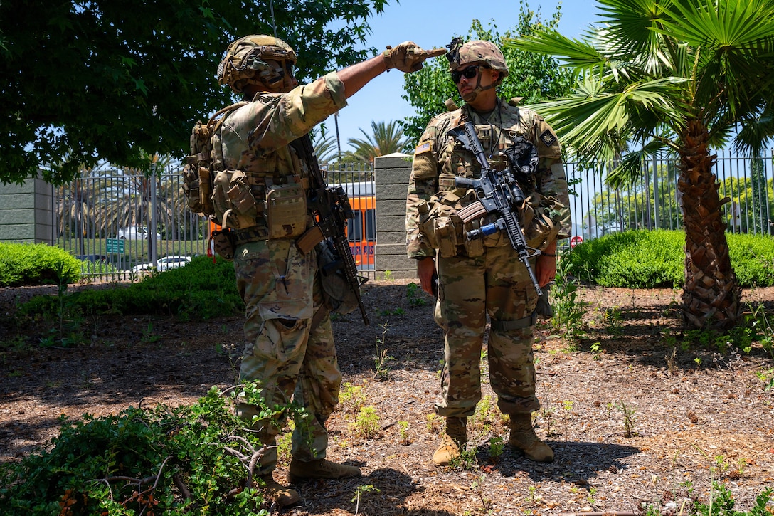 Two soldiers in combat gear, one pointing as the other looks on, stand outdoors on a sunny day with a gate, trees and vehicles in the background.