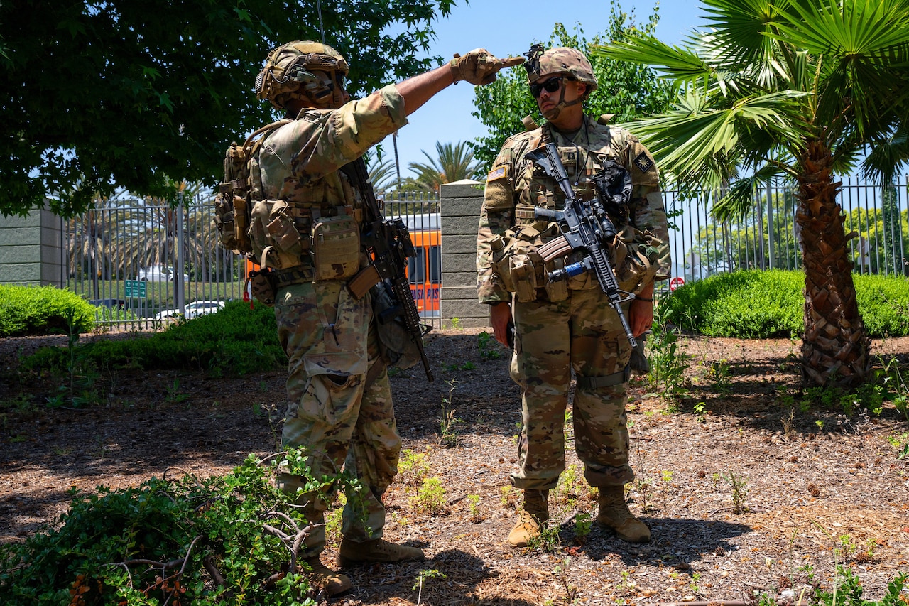 Two soldiers in combat gear, one pointing as the other looks on, stand outdoors on a sunny day with a gate, trees and vehicles in the background.