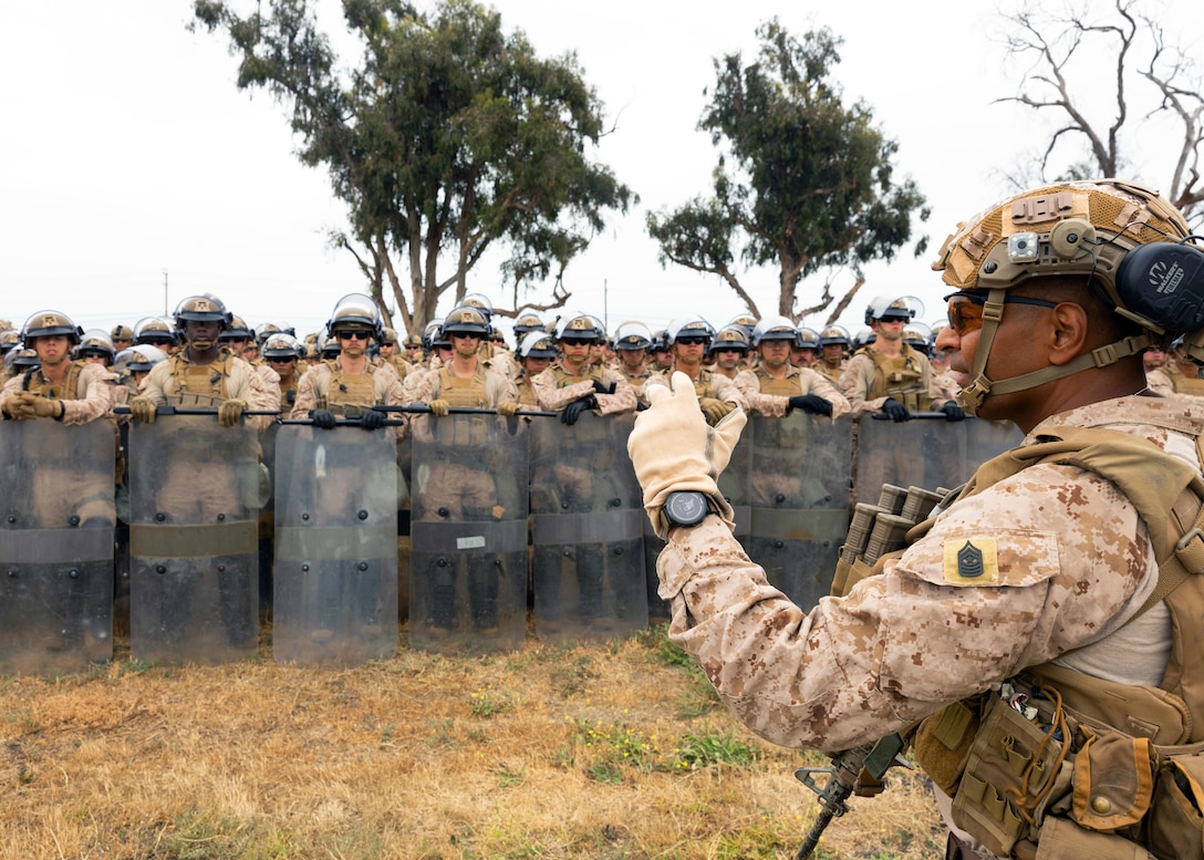 Marines in combat gear and holding shields listen as a Marine speaks to them on a grassy field with tress in the background.