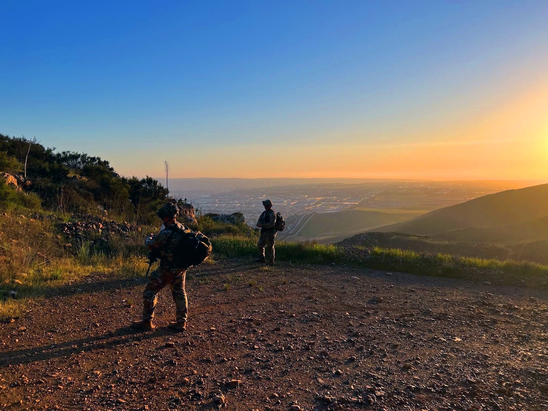 Two soldiers stand at an elevated area with a city seen in the hills below as the sun shines in the distance.
