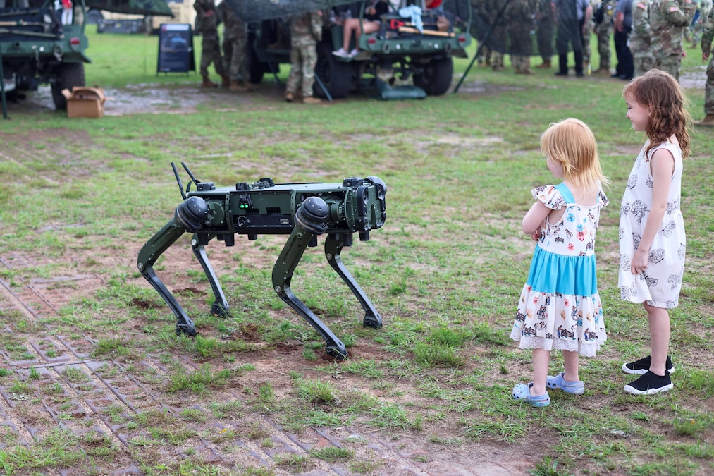 Two children look at a robot dog in a field with service members gathered in the background.