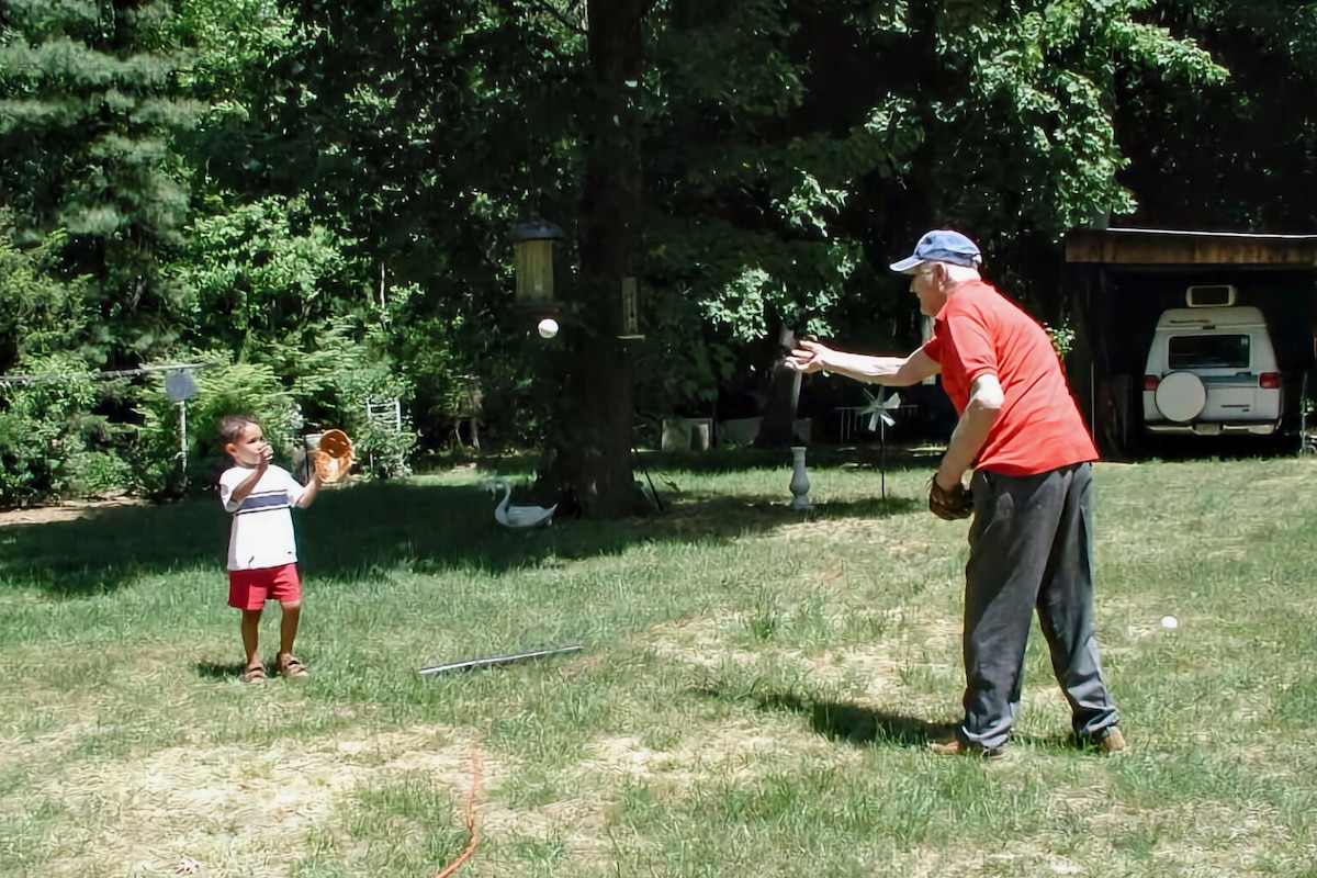 A boy wearing a baseball glove focuses on a baseball thrown by his grandfather in the front yard of a suburban home.