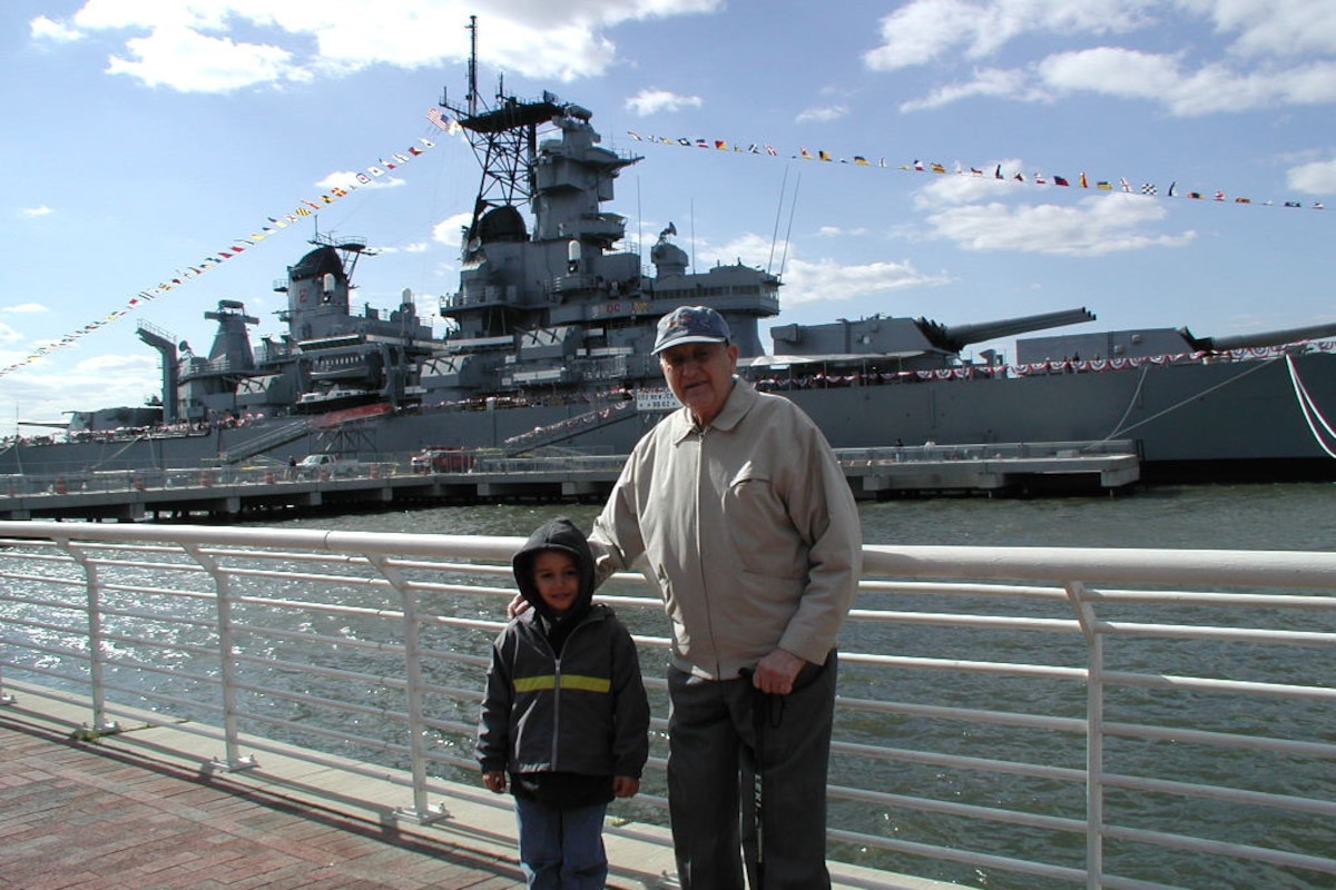 A boy wearing a hooded sweatshirt stands with his grandfather in front of a railing. A large ship adorned with pennants is docked in the background.