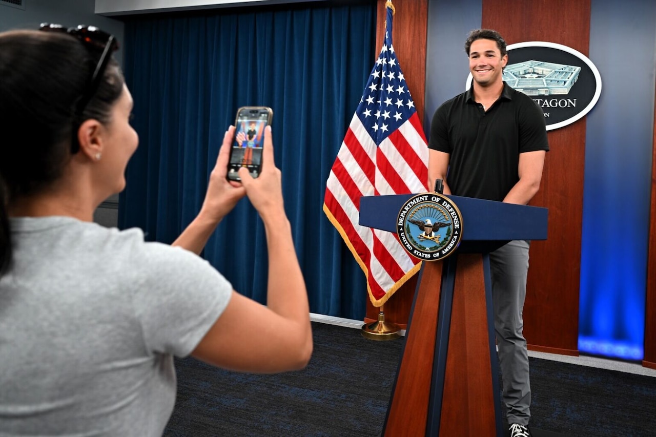 A man in casual attire stands in front of a podium as a woman takes his photograph with a mobile phone in the foreground.