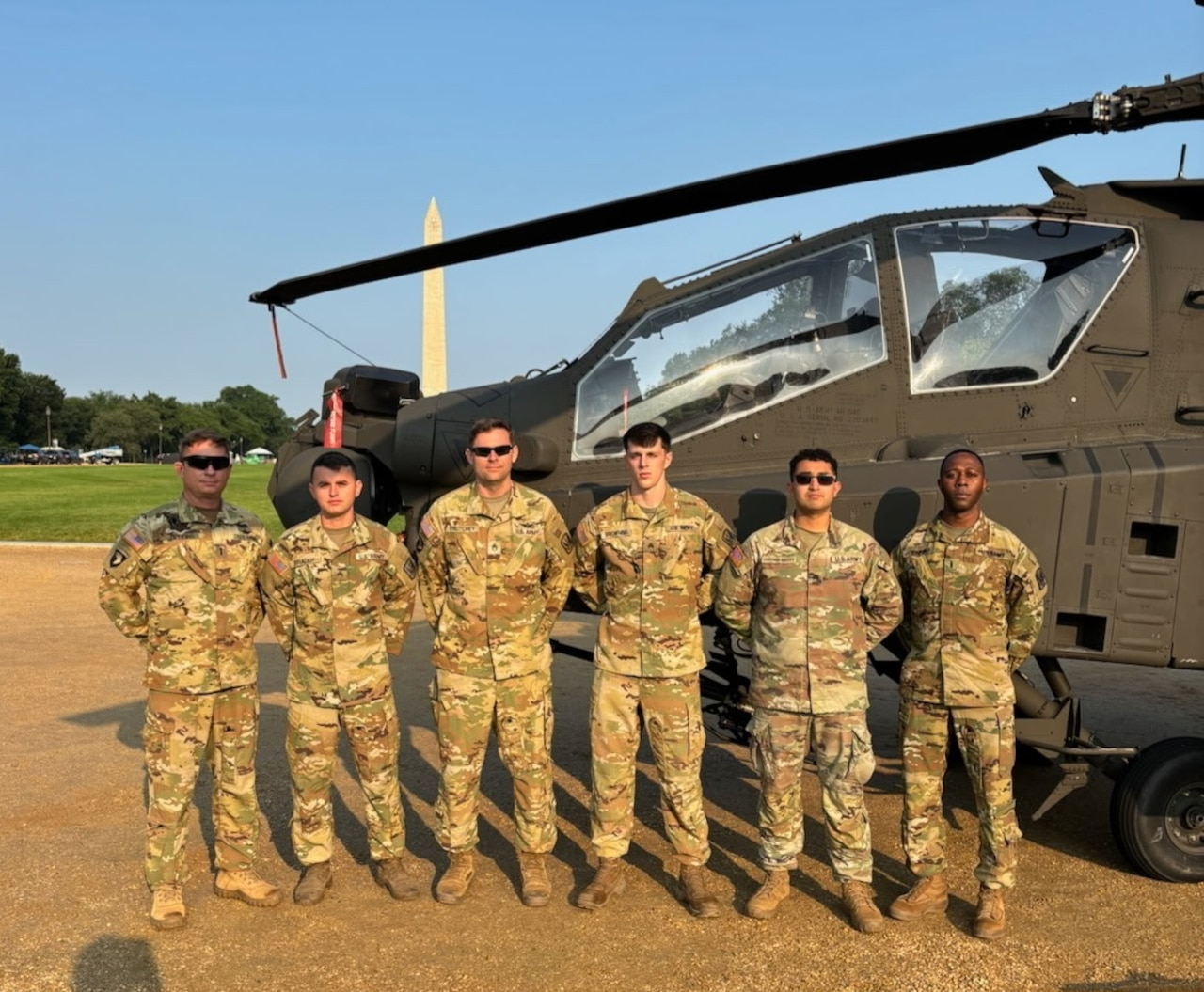 Six people wearing camouflage uniforms stand in front of a military helicopter. In the background is the Washington Monument.