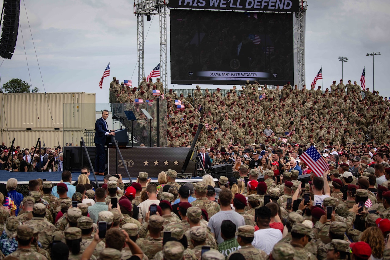 A man wearing business attire stands on a stage amidst hundreds of uniformed personnel who are standing on bleachers.