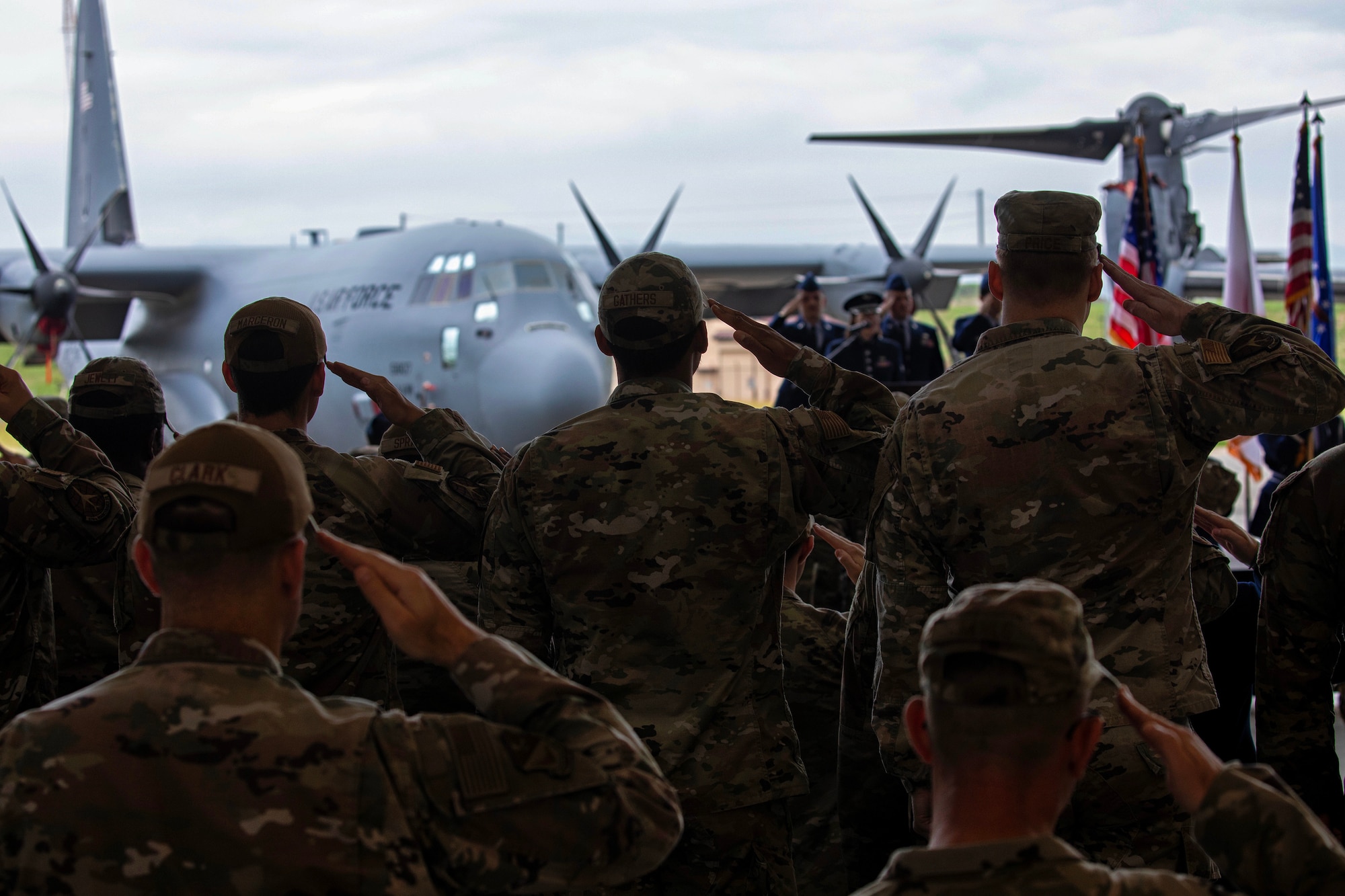 Airmen salute as the national anthem plays. A gray C-130J Super Hercules aircraft sits in the background.