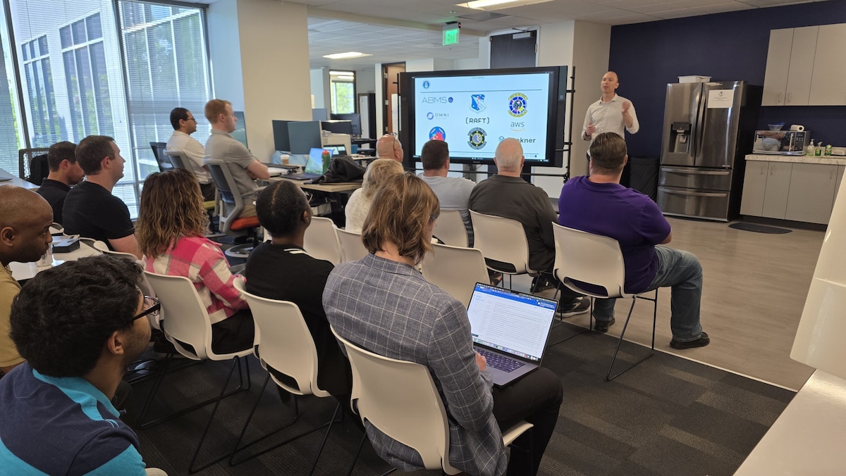 civilians sitting in chairs listening to a briefer standing by a large computer display