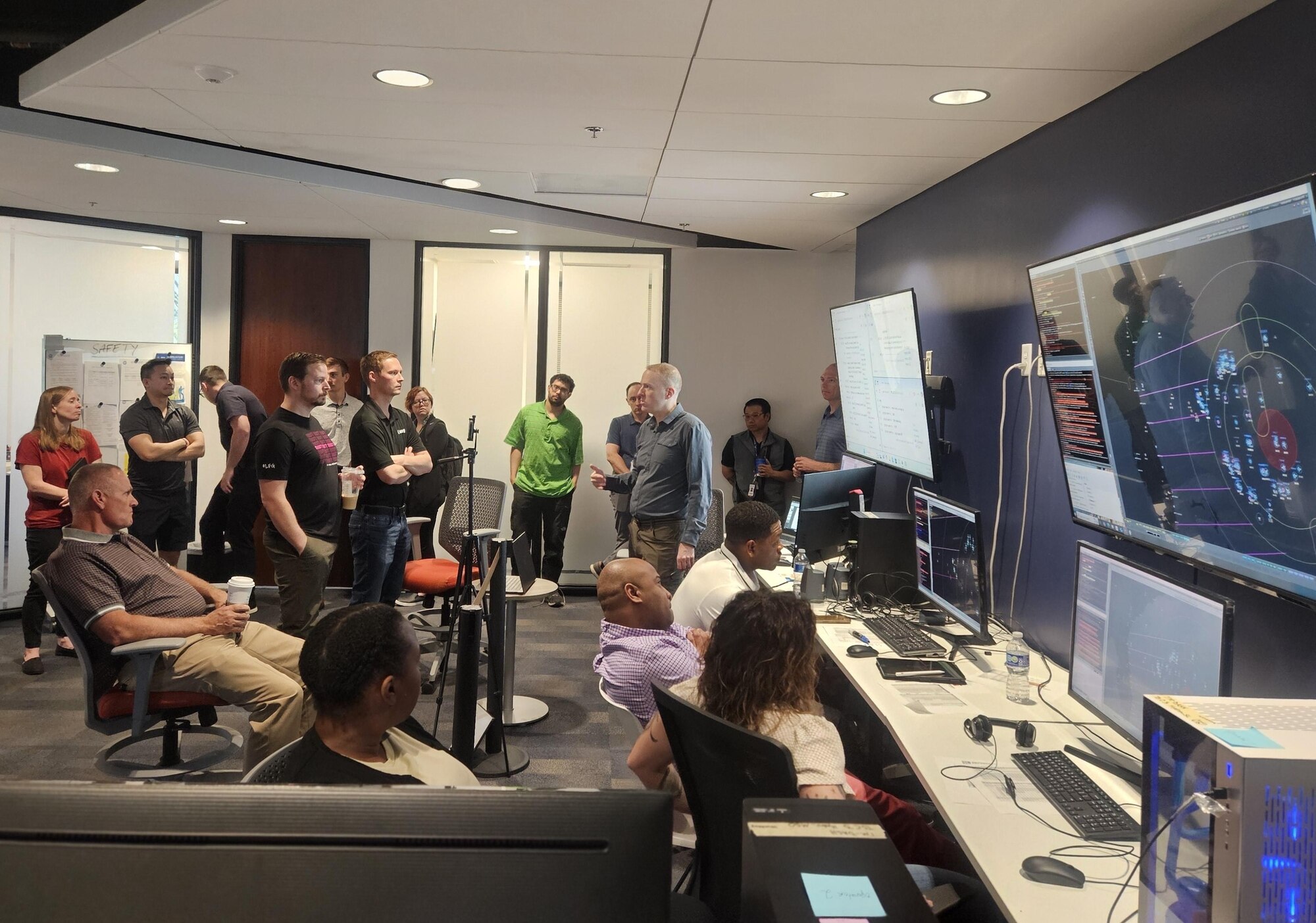 civilians sitting in chairs and standing listen to the leader standing, there are large computer screens mounted on the wall behind him and people working on computers