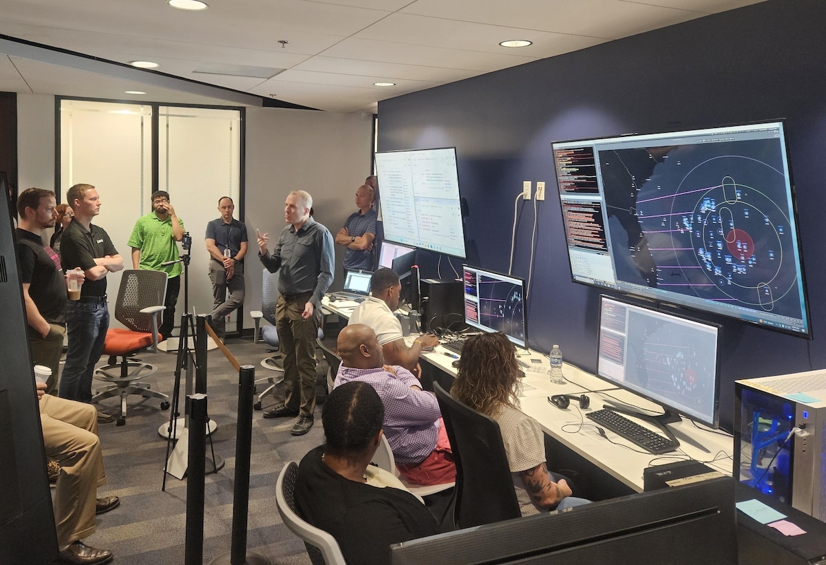 civilians sitting in chairs and standing listen to the leader standing, there are large computer screens mounted on the wall behind him and people working on computers