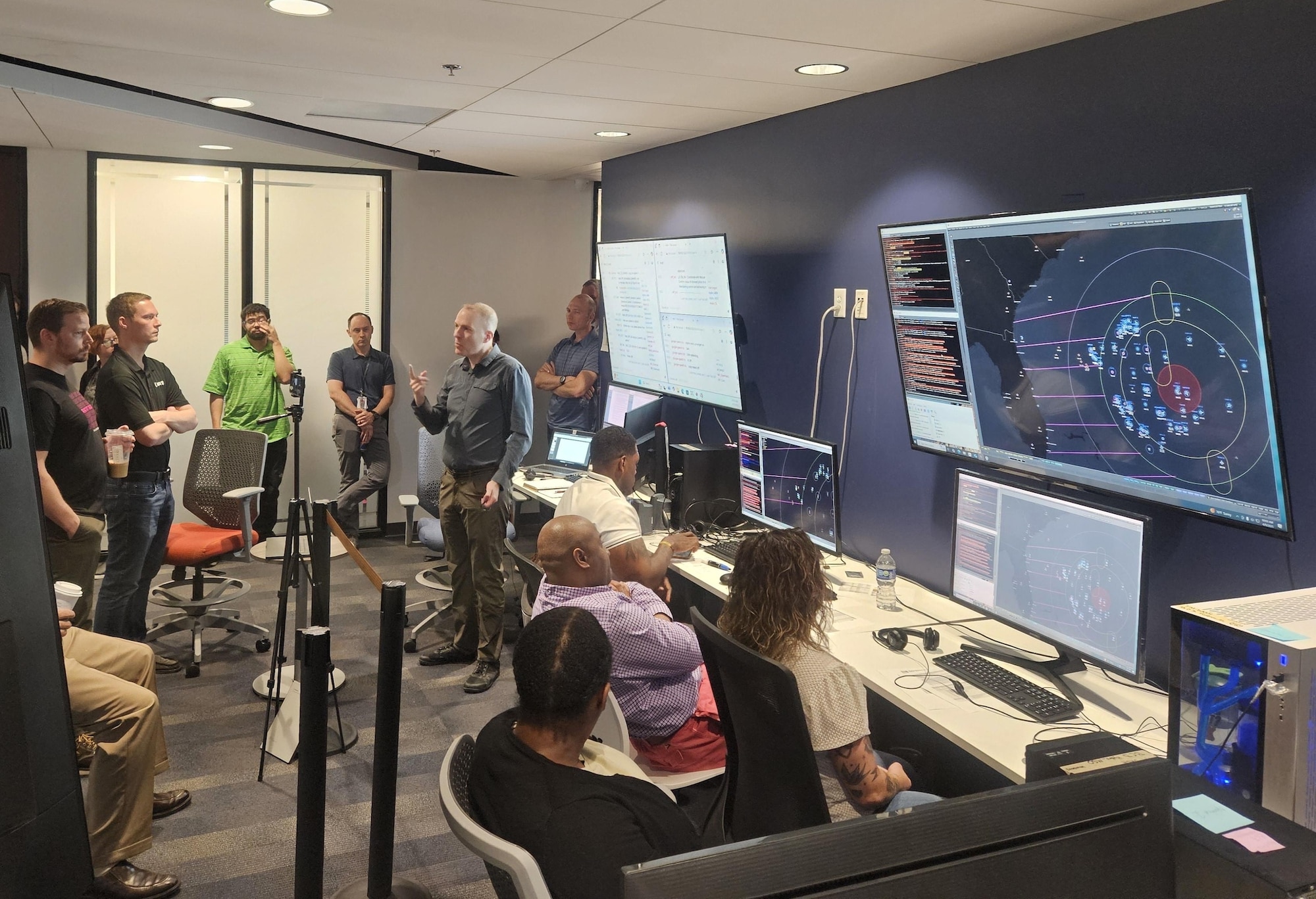 civilians sitting in chairs and standing listen to the leader standing, there are large computer screens mounted on the wall behind him and people working on computers