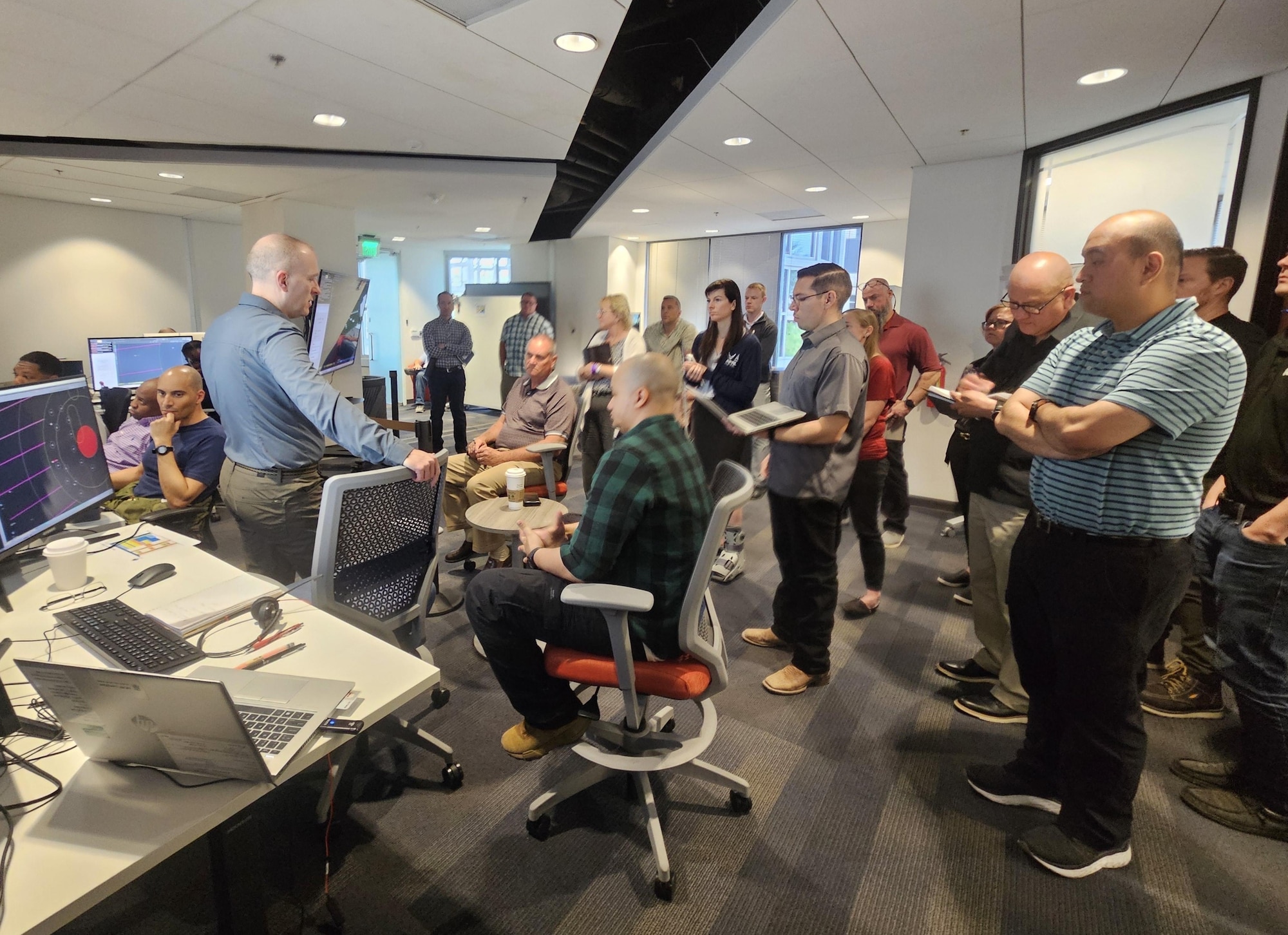 civilians sitting in chairs and standing listen to the leader standing, there is large computer screen mounted on the wall behind the people and people sitting working on computers in the room