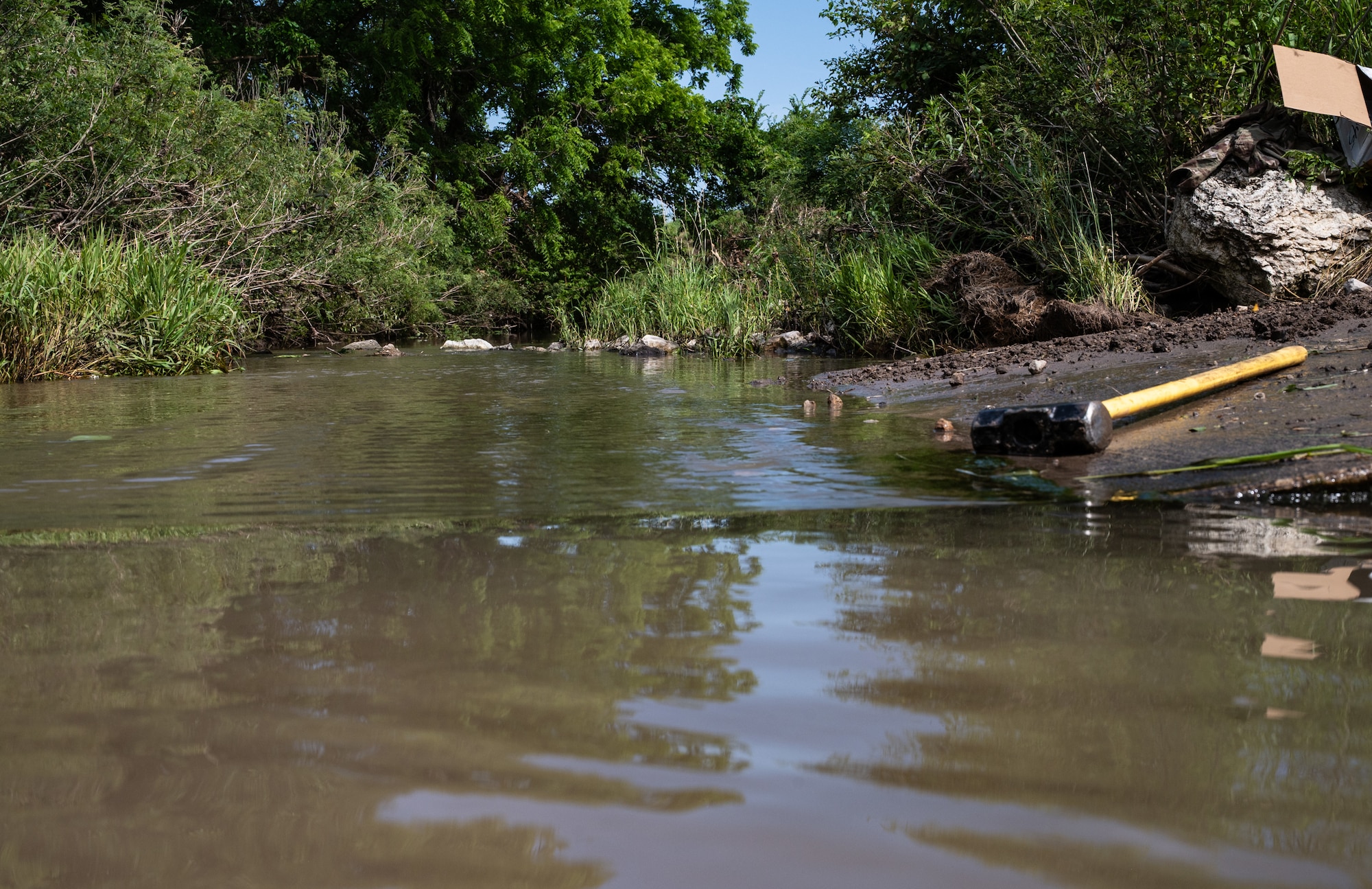 A sledgehammer lies on the bank of a creek during a bank revetment project June 11, 2025, at McConnell Air Force Base, Kansas. Airmen and civilians from the 22nd Civil Engineer Squadron, U.S. Fish and Wildlife Service, and Center for Environmental Management of Military Lands participated in the project that used trees attached to the bank to trap dirt and sediment so plant life can grow, allowing their roots to stabilize the bank and prevent erosion. (U.S. Air Force photo by Staff Sgt. Tryphena Mayhugh)