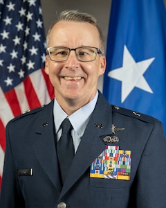 Official portrait of a main in dress uniform for the Air Force National Guard. He's wearing glasses, smiling, with an American flag and 1-star Air Force General flag in the background