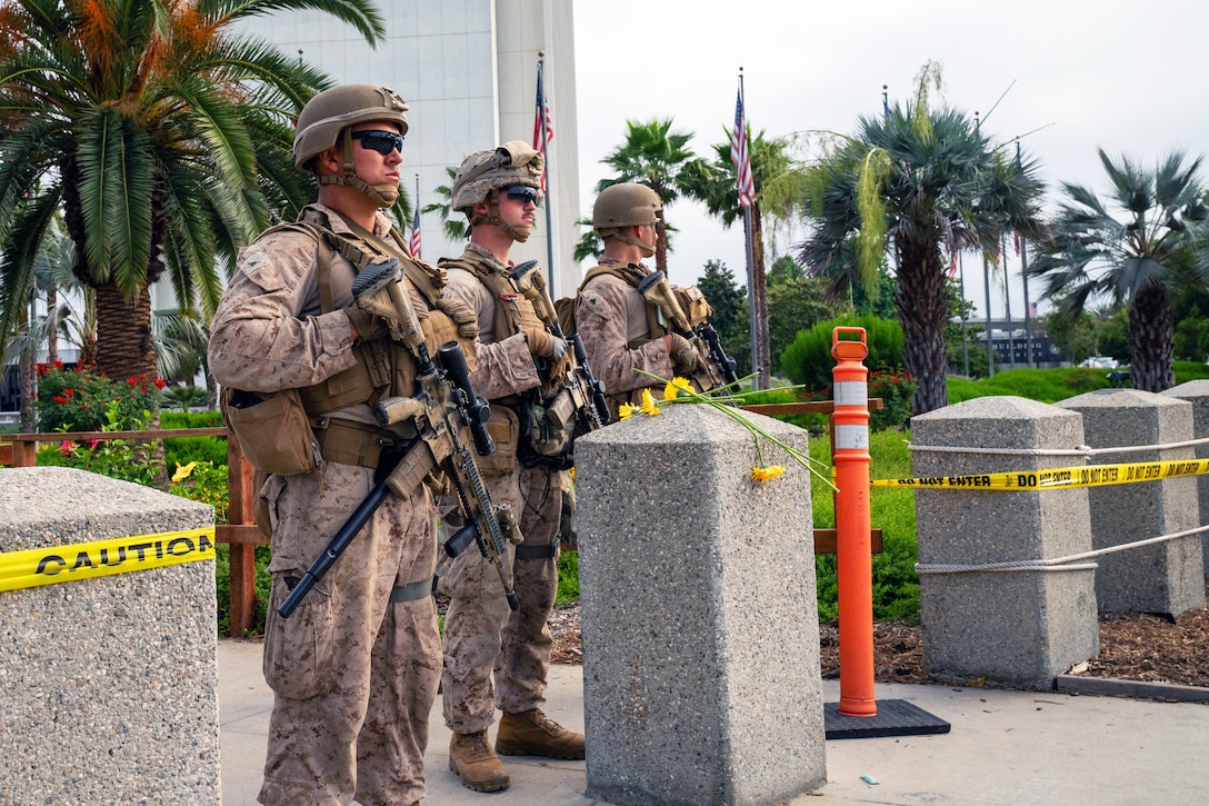 Three Marines stand at a checkpoint on a sidewalk with barriers and yellow caution tape outside a building with palm trees in the background.