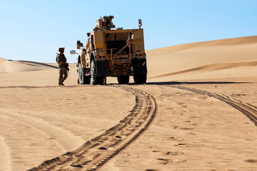 A soldier standing outside speaks to another soldier driving a tactical vehicle at a sandy area on a sunny day.