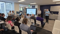 civilians sitting in chairs listening to two briefers standing by a large computer display