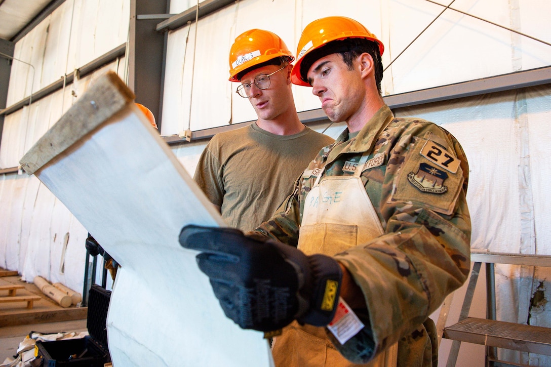 Two service members wearing construction hats look at a board while standing next to each other.