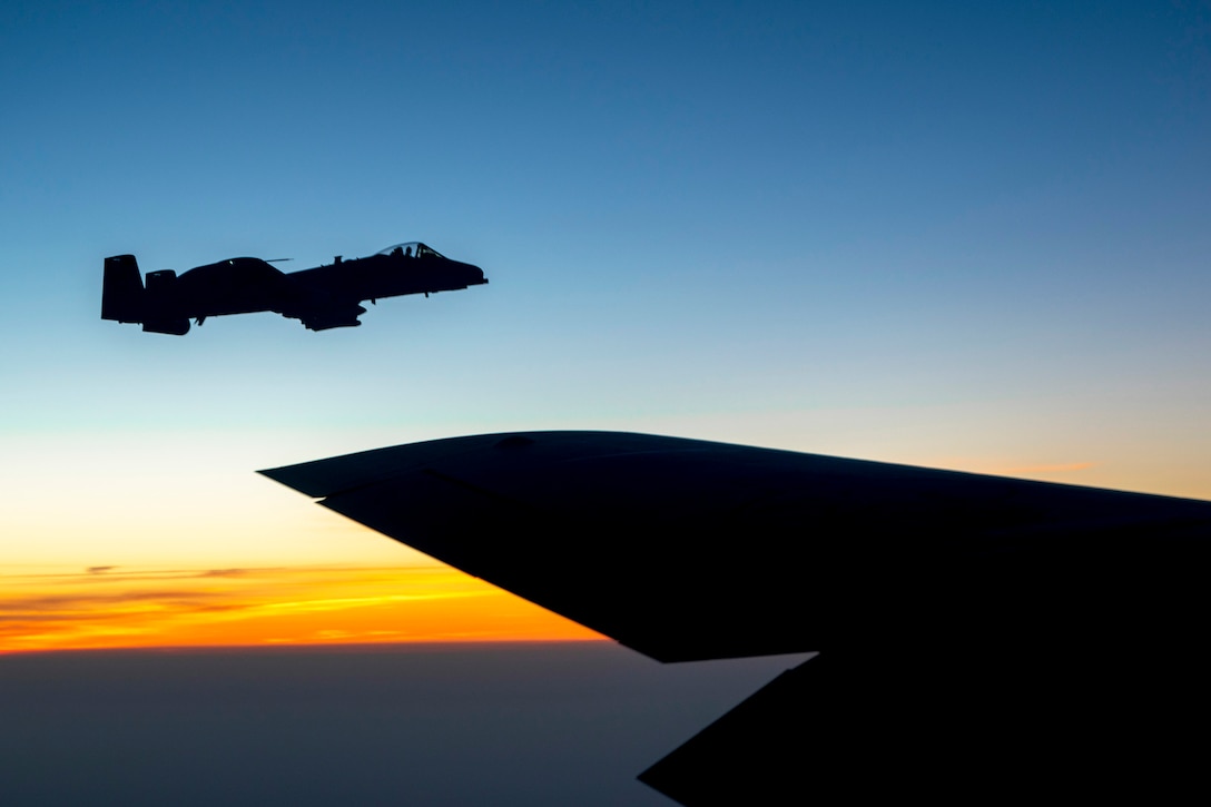 The wing of an aircraft is visible in the foreground as an aircraft in silhouette flies in a blue and orange-streaked sky.