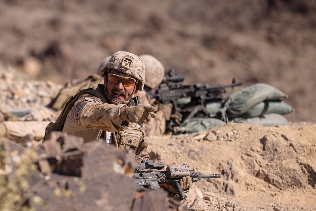 A Marine in tactical gear points while kneeling in a desert-like area as fellow Marines aim weapons in the background.