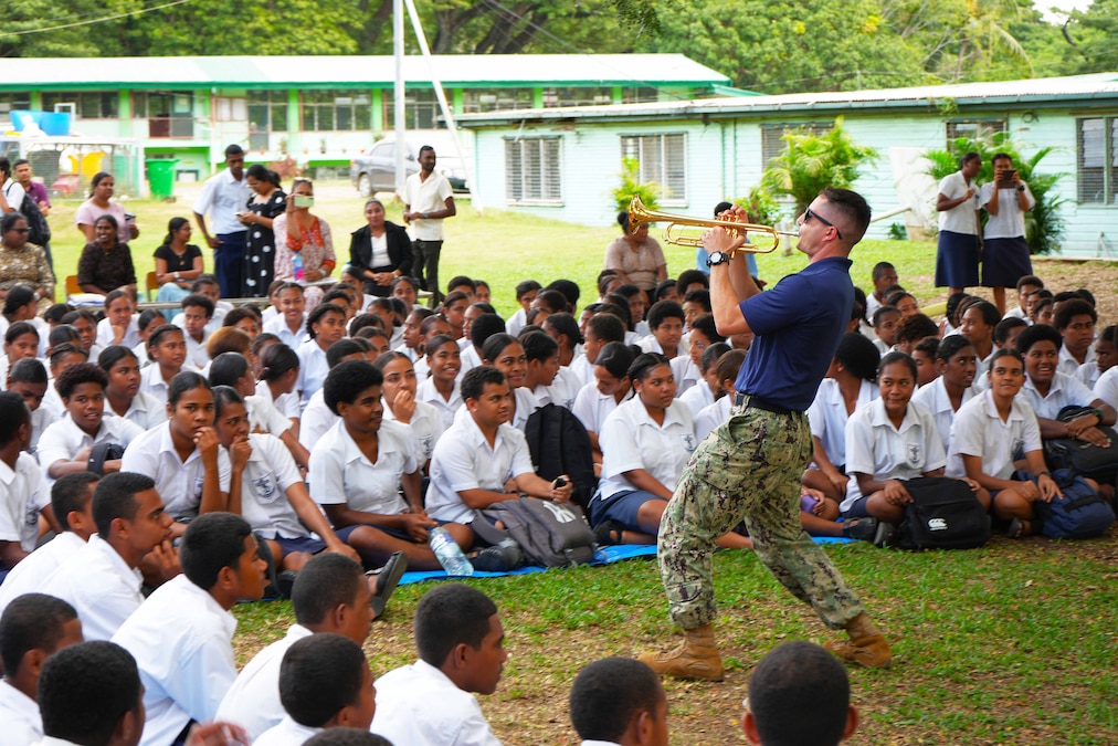 A sailor plays the trumpet in front of dozens of seated high school students outside of a green building.