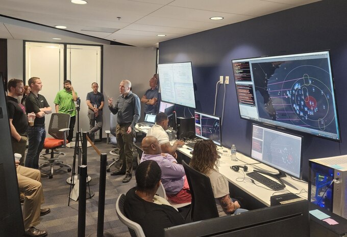 civilians sitting in chairs and standing listen to the leader standing, there are large computer screens mounted on the wall behind him and people working on computers