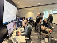 people in civilian clothes sit working on computers while others stand or sit behind them watching