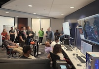 civilians sitting in chairs and standing listen to the leader standing, there are large computer screens mounted on the wall behind him and people working on computers