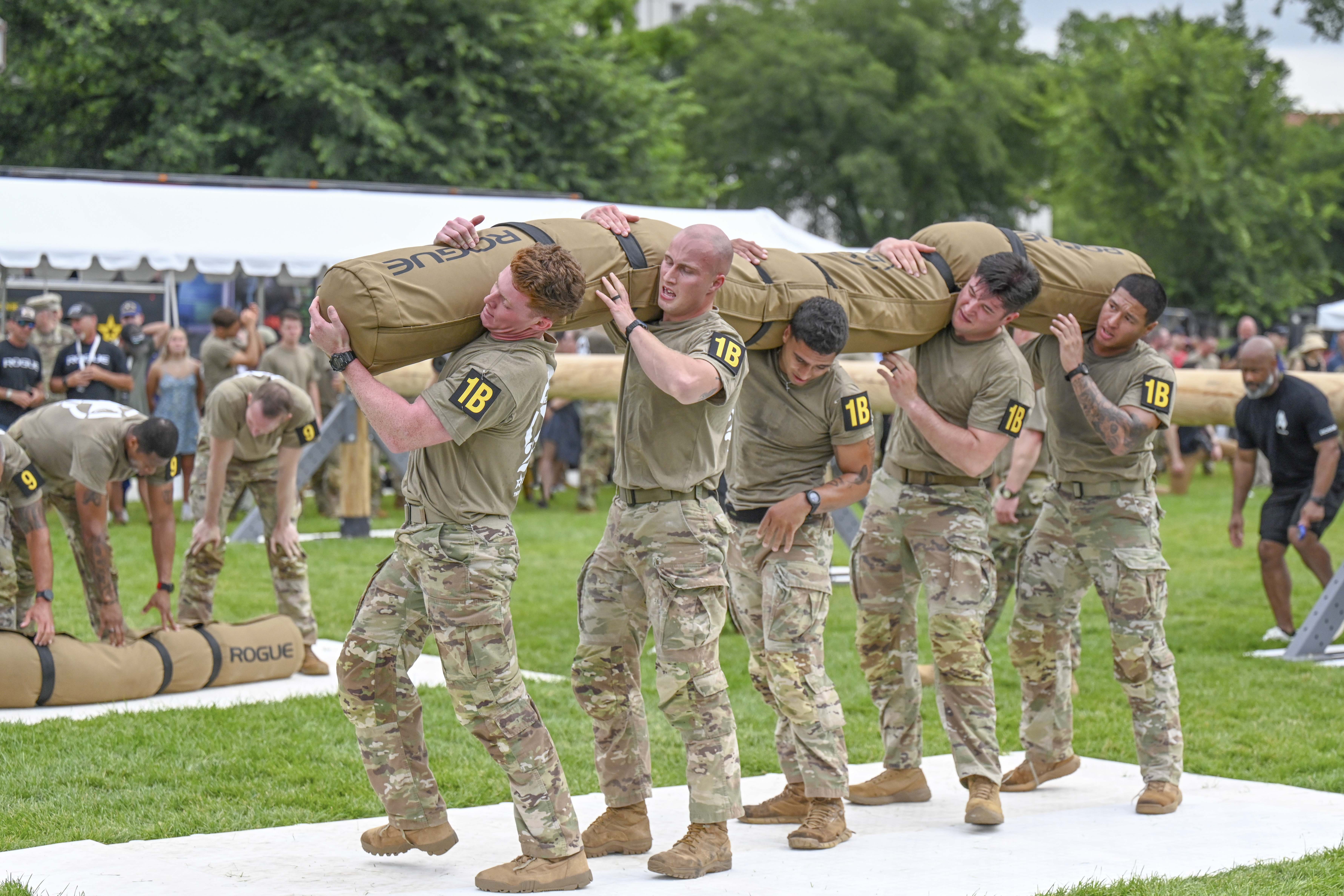 Five soldiers lift a weighted bag on a grassy area as people watch on the sideline.
