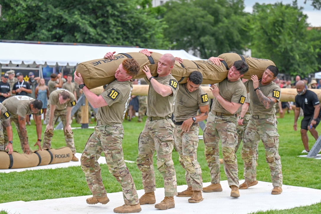 Five soldiers lift a weighted bag in a field as people watch on the sideline.