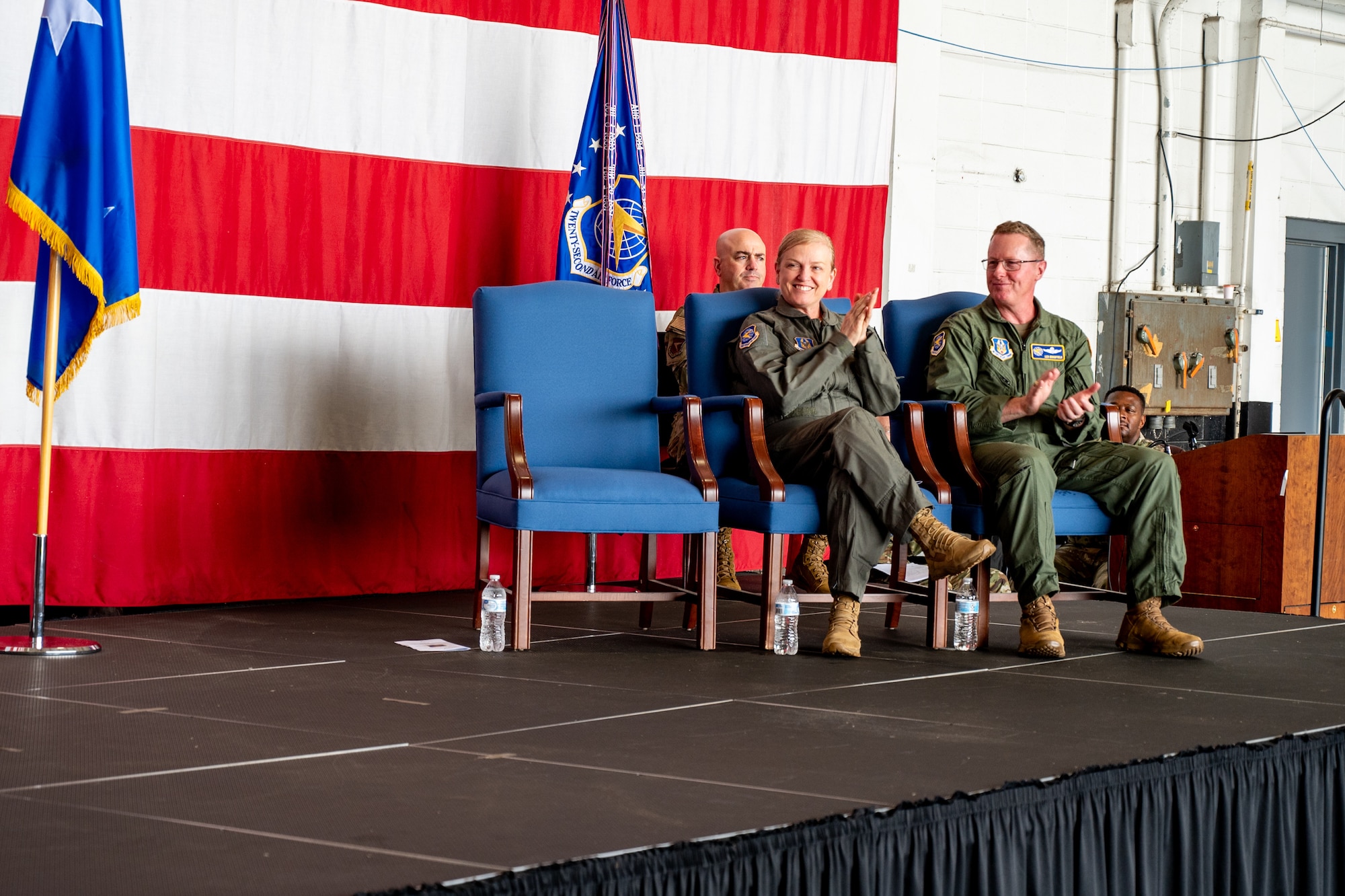 a man and woman in military uniforms clap while sitting in blue chairs on stage