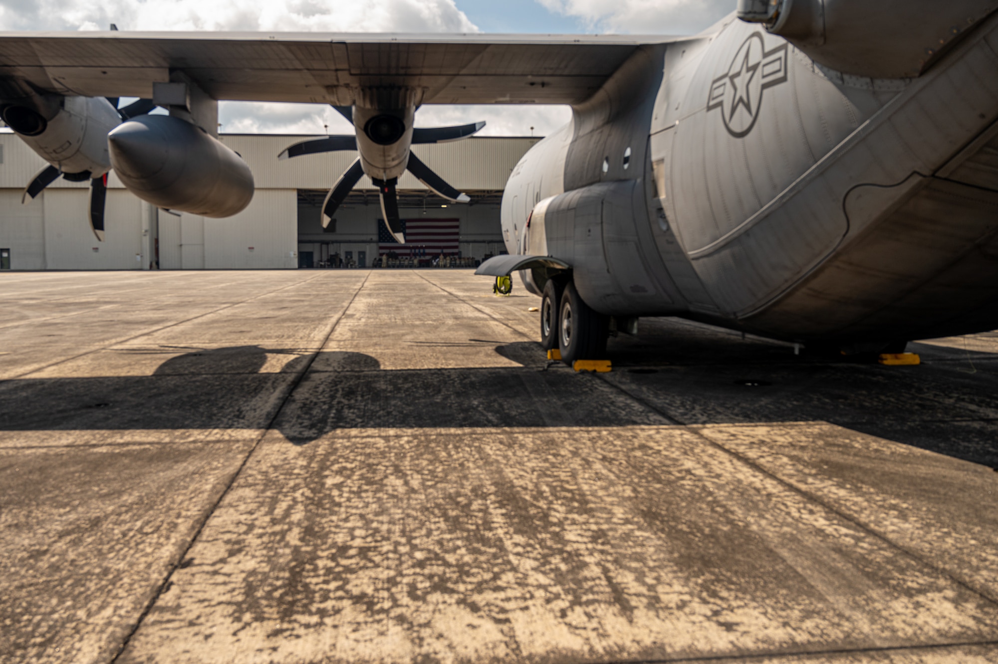 a C-130 aircraft sits on a ramp in front of open hangar doors