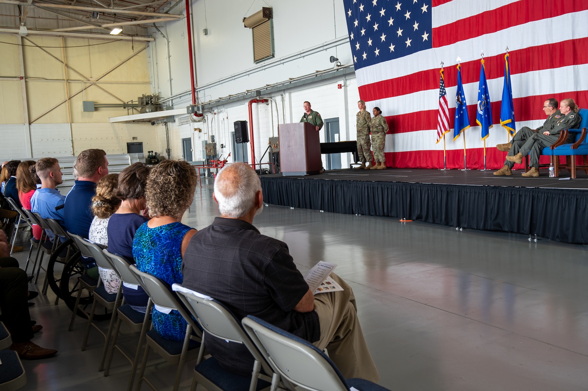 people sit in chairs in an audience while a man in a military uniform stands on stage behind a lectern and delivers a speech
