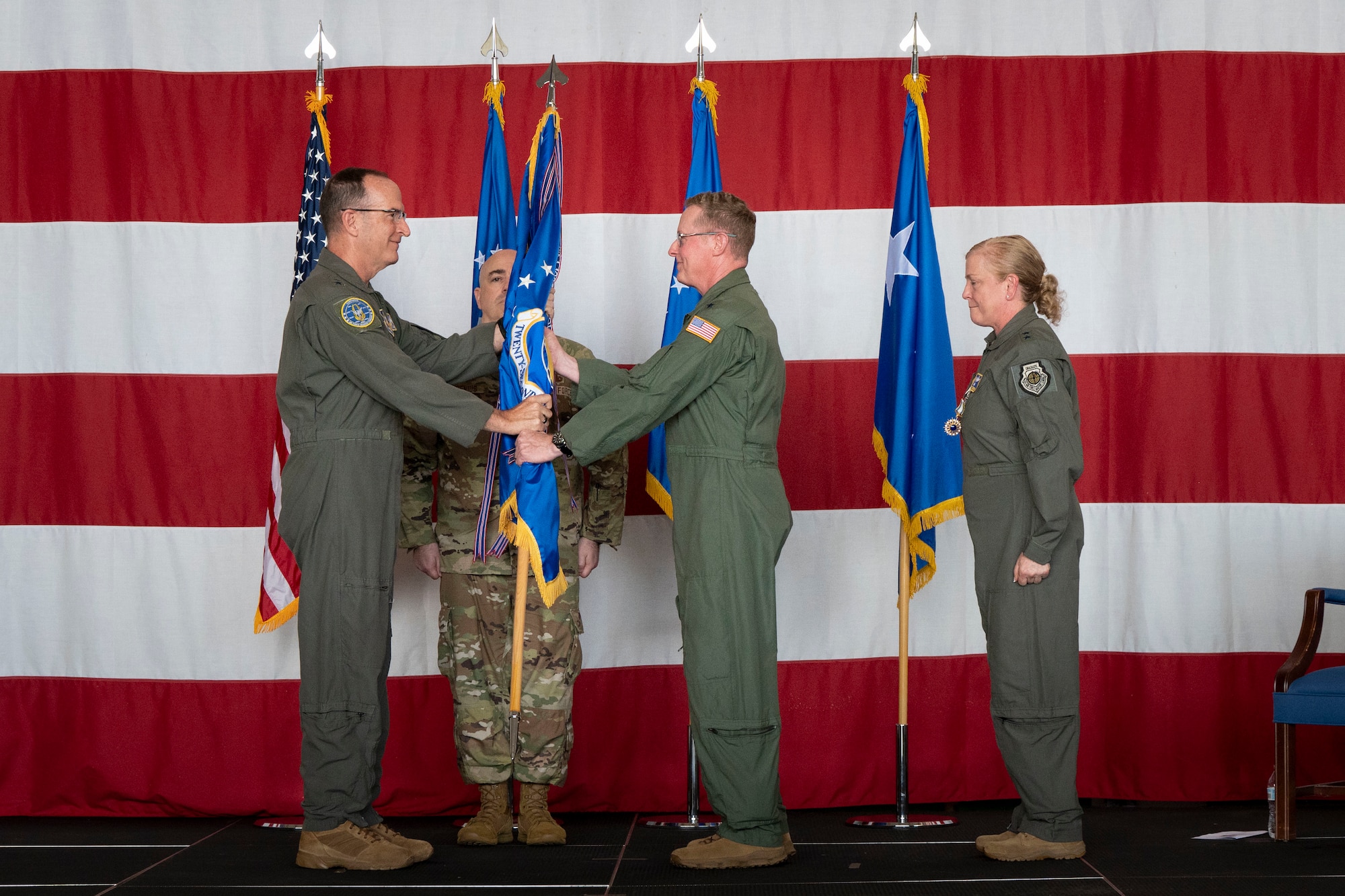 two men in military uniforms face each other holding a guidon on stage in front of an American flag while two other people in uniform look on