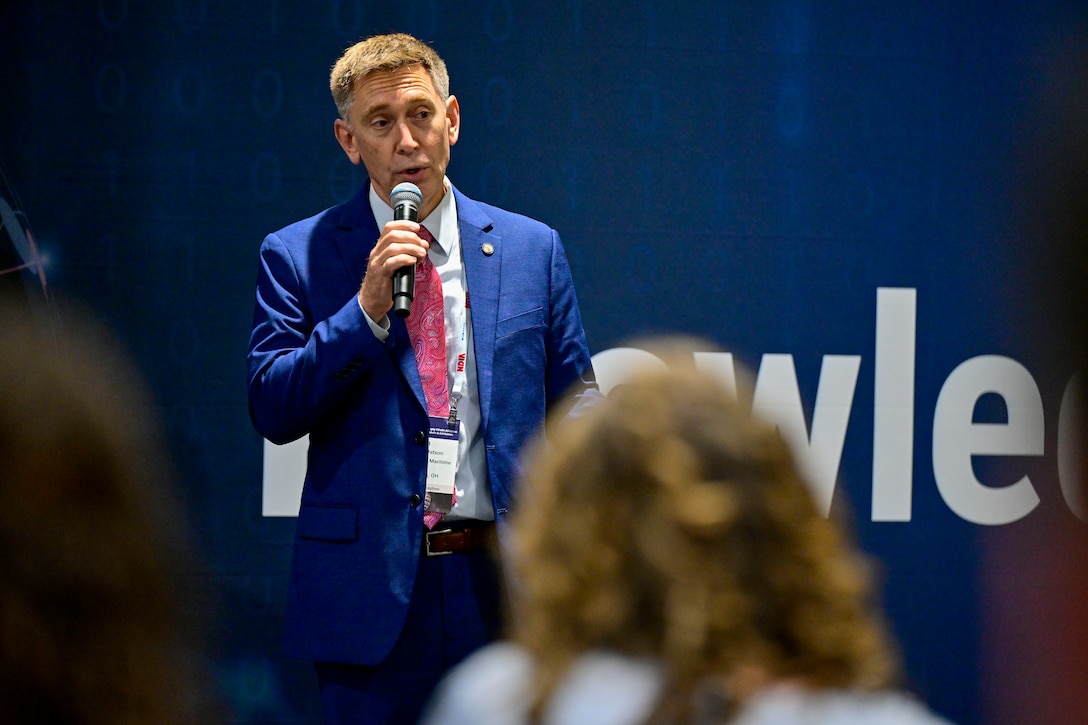 Close up of man in blue suit speaking, holding a microphone. Blue background with white letters.