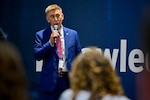 Close up of man in blue suit speaking, holding a microphone. Blue background with white letters.