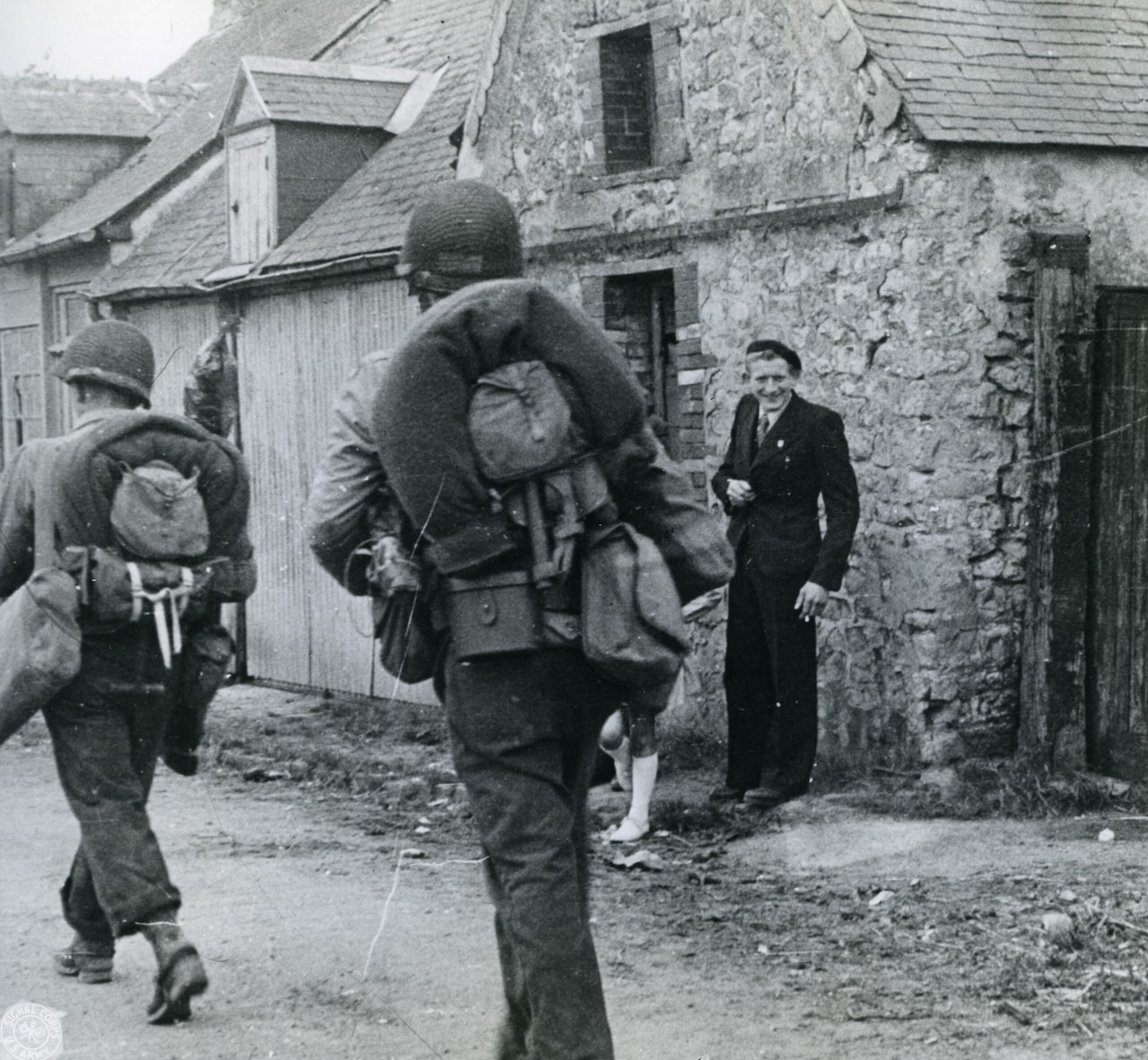 Two men in military uniforms carrying rucksacks walk along a street. A man, wearing a suit and standing by a home, smiles at them.