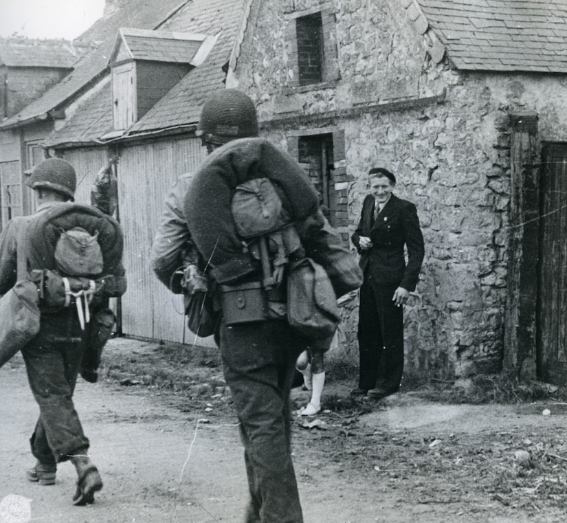 Two men in military uniforms carrying rucksacks walk along a street. A man, wearing a suit and standing by a home, smiles at them.