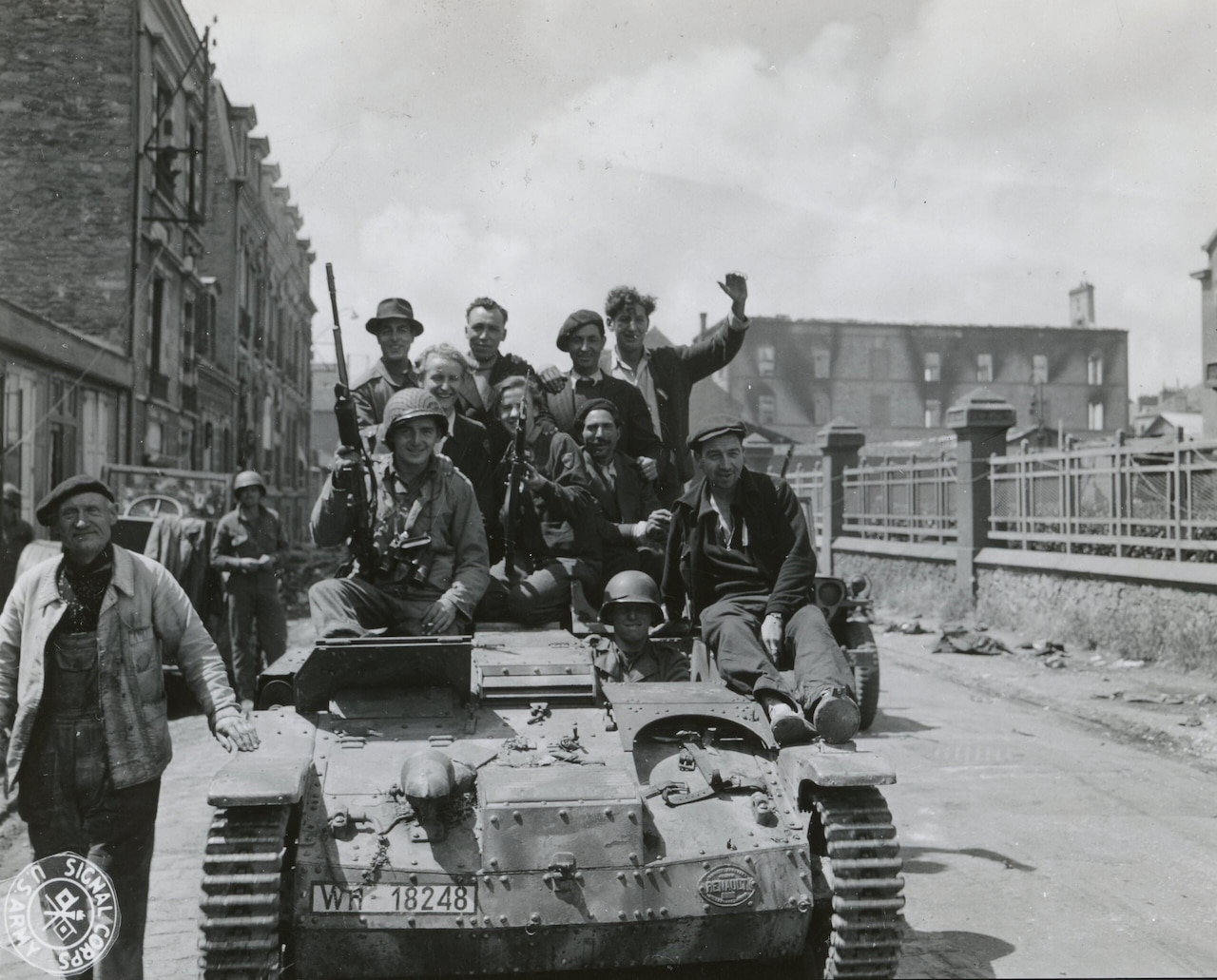 Two men in military uniforms and several men in civilian attire ride on top of a tank as it moves through a street. Two men are holding guns, and another man in civilian clothes is standing beside the tank.