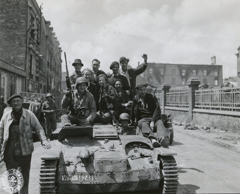 Two men in military uniforms and several men in civilian attire ride on top of a tank as it moves through a street. Two men are holding guns, and another man in civilian clothes is standing beside the tank.