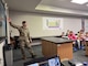 Man in uniform pointing to a screen while lecturing to students.