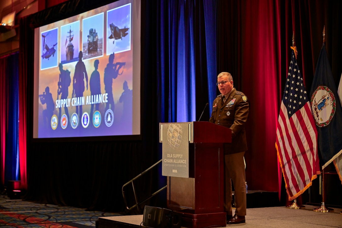 An Army lieutenant general speaks at a podium with military flags in the background and a screen with a PowerPoint slide.