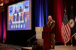 An Army lieutenant general speaks at a podium with military flags in the background and a screen with a PowerPoint slide.